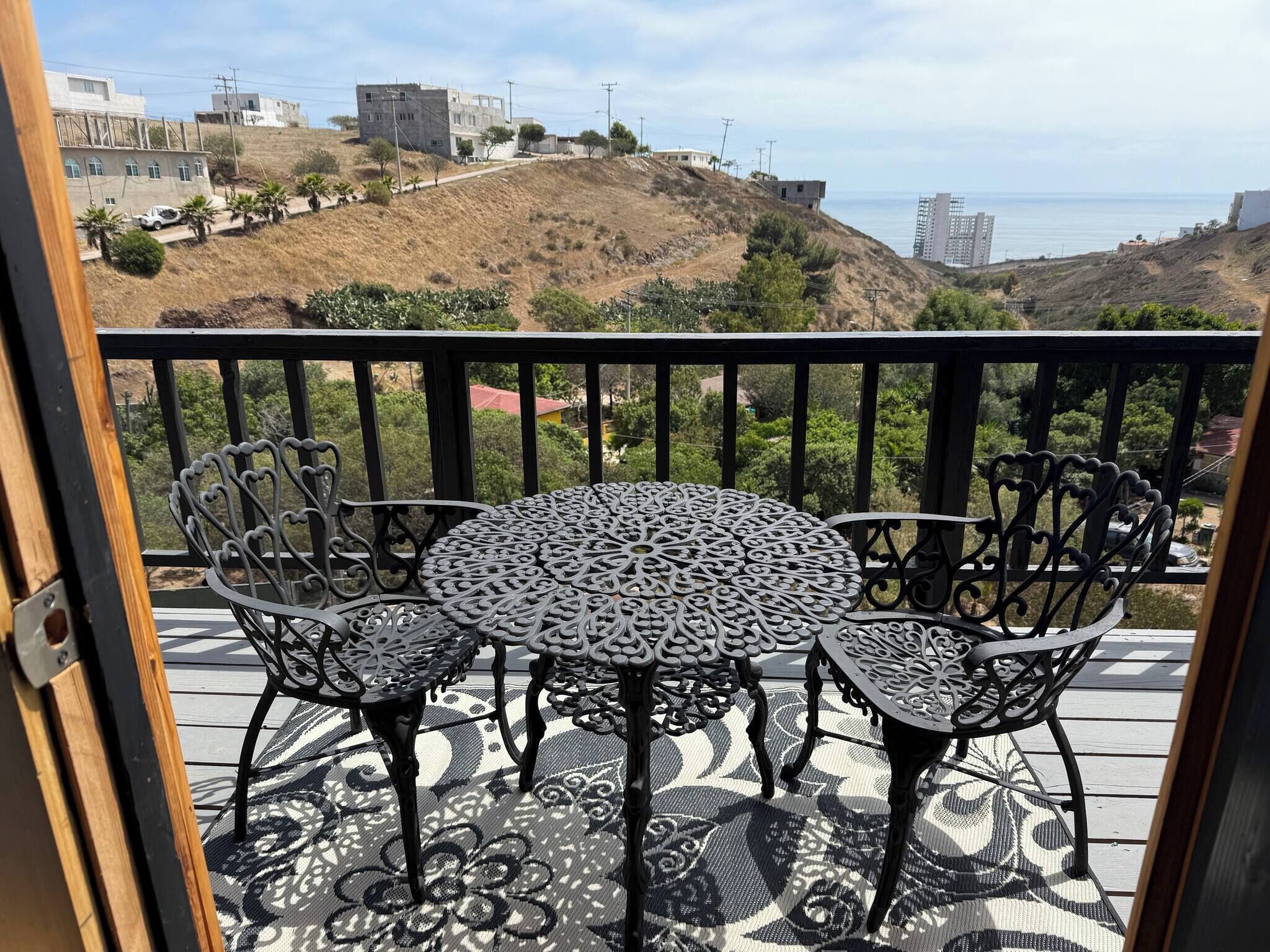 Ein Balkon mit rundem Tisch und vier Stühlen, der auf eine Hügelkuppe mit Gebäuden und Meerblick blickt.