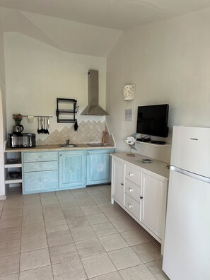 Eine kompakte Küche mit Mikrowelle, Wasserkocher und Toaster auf der Arbeitsplatte, einem Herd und einem Spülbecken. Ein weißer Kühlschrank und ein Fernseher auf einem Lowboard stehen dort.
