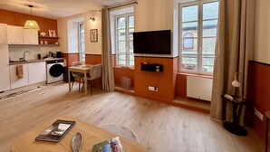 A modern kitchen with a washing machine, a dining area with a table and chairs, a television mounted on the wall, and large windows with curtains.