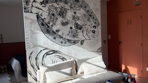 A modern bedroom with a large abstract wall art, a grey sofa, a wooden coffee table with various items, and a hanging light fixture.