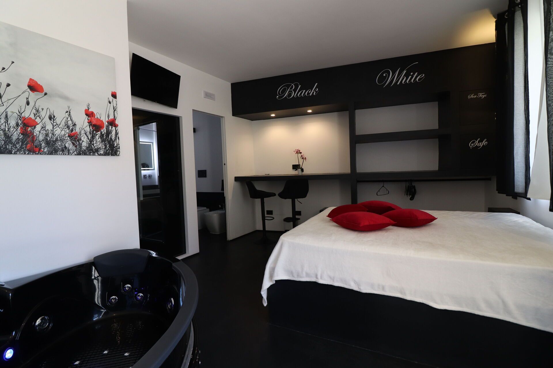 A modern hotel room with a black and white color scheme, a bed with red pillows, a desk with two chairs, and a painting on the wall.
