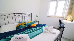 Una cama bien hecha con almohadas coloridas y una manta color turquesa, una mesita de noche con lámpara, una silla y una ventana con cortinas.