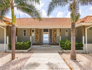 A building with a tiled roof, a covered walkway, and palm trees. - Kas di Sono Pool & Jacuzzi Private Beach (Sint Willibrordus)