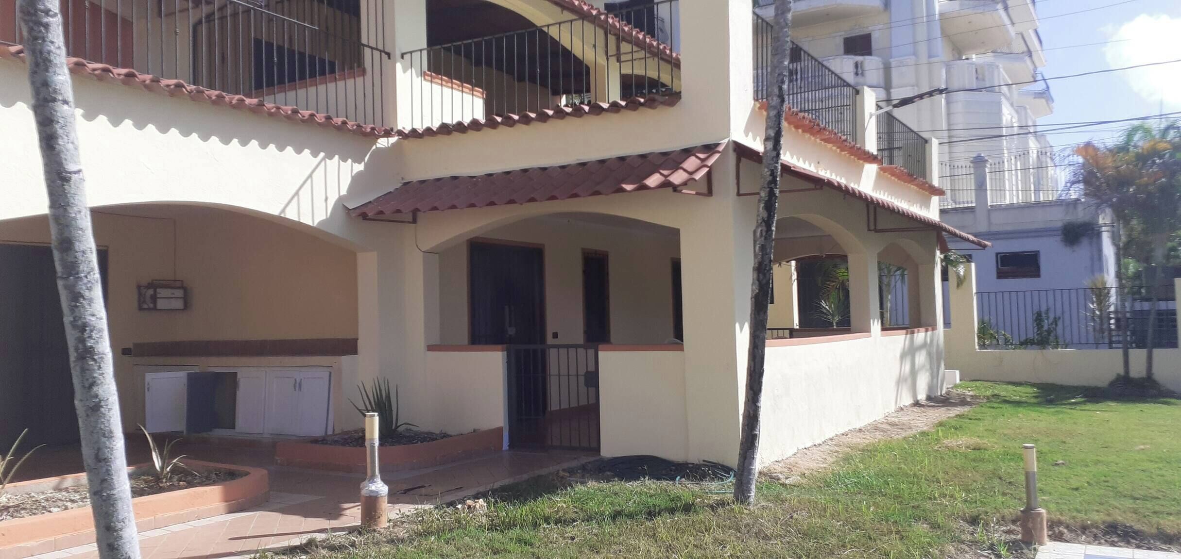 A two-story building with a tiled roof, a covered porch, and a small garden area with a tree and a bottle of water.
