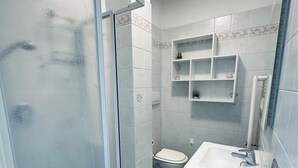 Um banheiro com box de vidro, vaso sanitário, pia com armário azul e prateleira na parede com artigos de higiene pessoal.