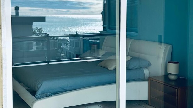 Quarto com cama, mesa de cabeceira e vista para o oceano através de uma porta de vidro.
