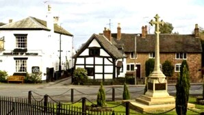 A traditional pub with a white facade and black timber framing, a stone monument in the foreground, and a black metal fence surrounding the area.