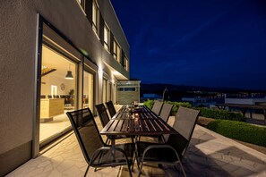 An outdoor patio with a wooden table and chairs, a glass door leading to an interior space, and a view of the city at night. - Charming Villa With Heated Pool (Posedarje)