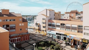 Un pueblo costero con noria, edificios frente al mar y diversas tiendas, incluyendo un restaurante de sushi.