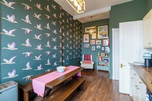 Een kamer met een houten bankje, een tafel met een roze tafelkleed, een witte stoel en een muur met een vogelmotief.