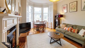 A living room with a fireplace, a green sofa, a dining table with chairs, and a large window. - Gorgeous 2BDR with Unique Character in Edinburgh (Edinburgh)