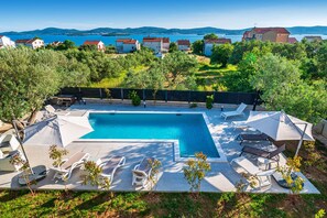 Ein Pool mit Liegestühlen und Sonnenschirmen, umgeben von Grünflächen und Häusern im Hintergrund.