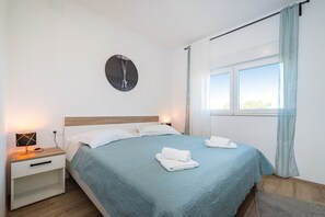 Ein ordentlich bezogenes Bett mit blauer Tagesdecke, weißen Kissen und sauber gefalteten Handtüchern. Ein Nachttisch mit Lampe und einer Wanduhr sind vorhanden.