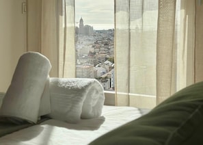 Ein Hotelzimmer mit Bett, einem Fenster mit transparenten Vorhängen und Blick auf eine Stadtlandschaft.