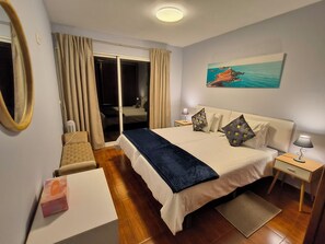 A hotel room with a bed, a desk, a chair, a nightstand, a lamp, a painting, and a sliding door to the balcony. - Golden Sun Refuge (Estreito da Calheta)