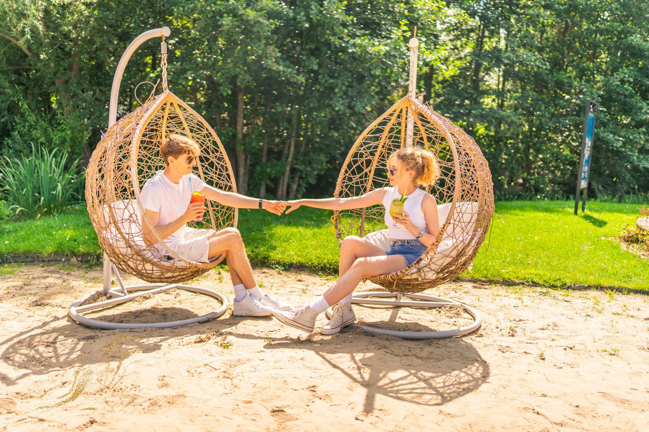 Two people sitting in wicker swing chairs on a sandy surface with greenery in the background.