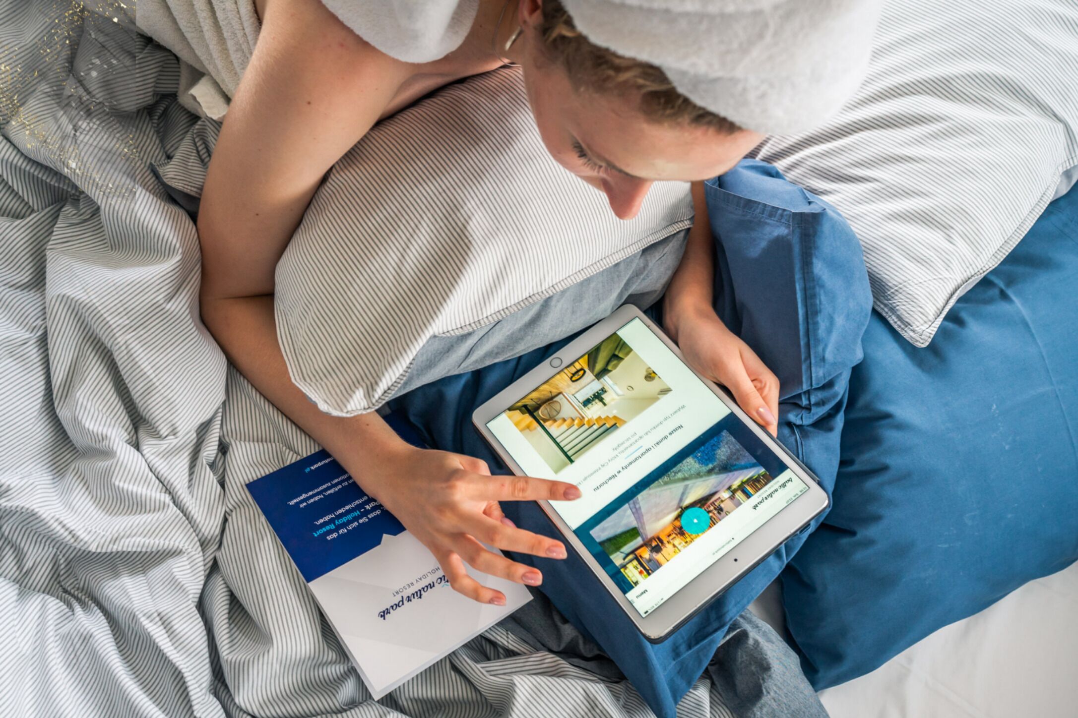 Una persona acostada en una cama con una tableta mostrando la página web de un hotel, un folleto y una almohada.