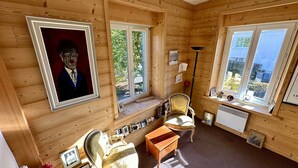 A cozy cabin interior with wooden walls, a framed portrait, a chair, a small table, and a radiator. - Sunny Sanctuary by the Town Center (Mont-Dore)