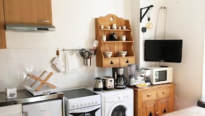 A compact kitchen with a stove, oven, washing machine, microwave, and a wooden shelf with cups and mugs. - Le Salers -Le Mont Dore 2pers (Mont-Dore)