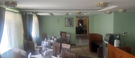 A dining area with tables set for service, wooden chairs, a kitchen area with appliances, and a view into the dining room.