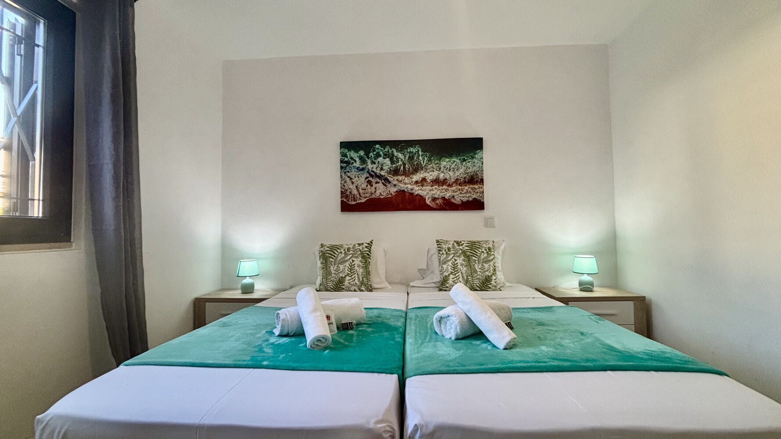 A hotel room with two beds, each with a green bedspread and white linens. There are two nightstands with lamps, a painting on the wall, and a window with curtains.
