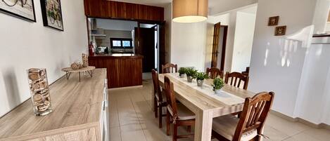 A dining area with a wooden table and chairs, a kitchen in the background, and a hanging light fixture.