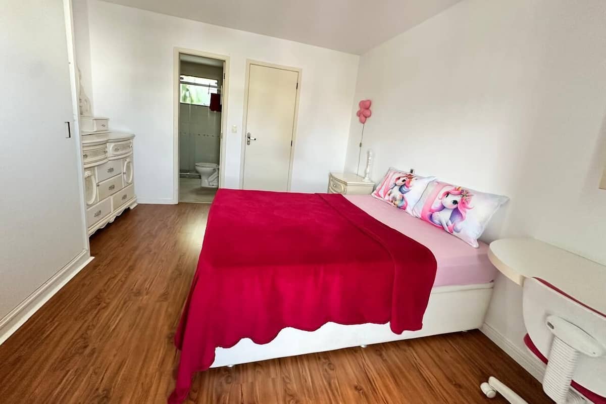 Una camera da letto con pavimento in legno, un letto con coperta rossa, un comodino con cuscino rosa e una cassettiera bianca.