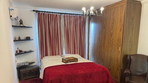 Een slaapkamer met een bed, een dressoir, een bureau, een stoel, een plank met decoratieve objecten en een gordijn.
