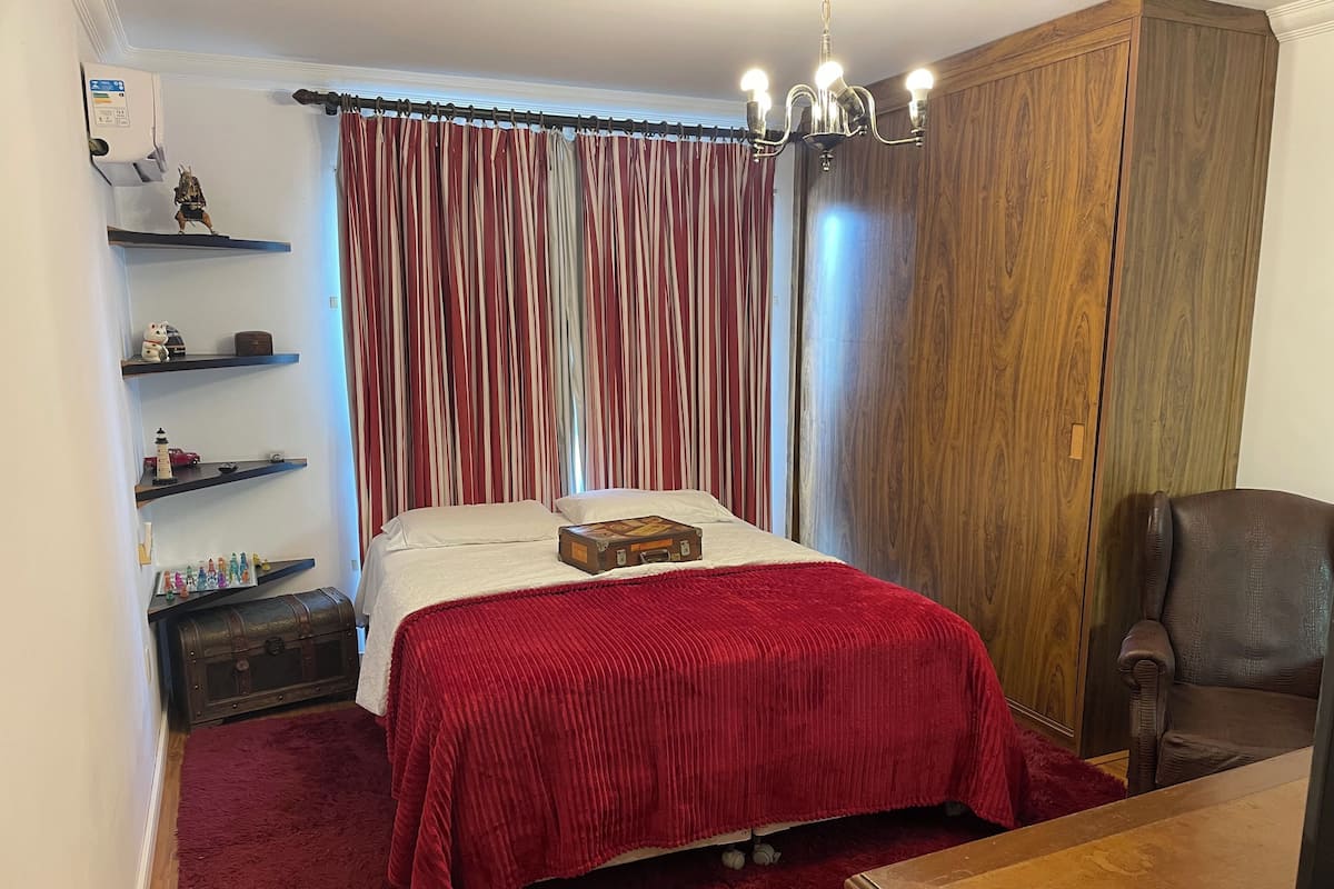 Una camera da letto con un letto, una cassettiera, una scrivania, una sedia, uno scaffale con oggetti decorativi e una tenda.
