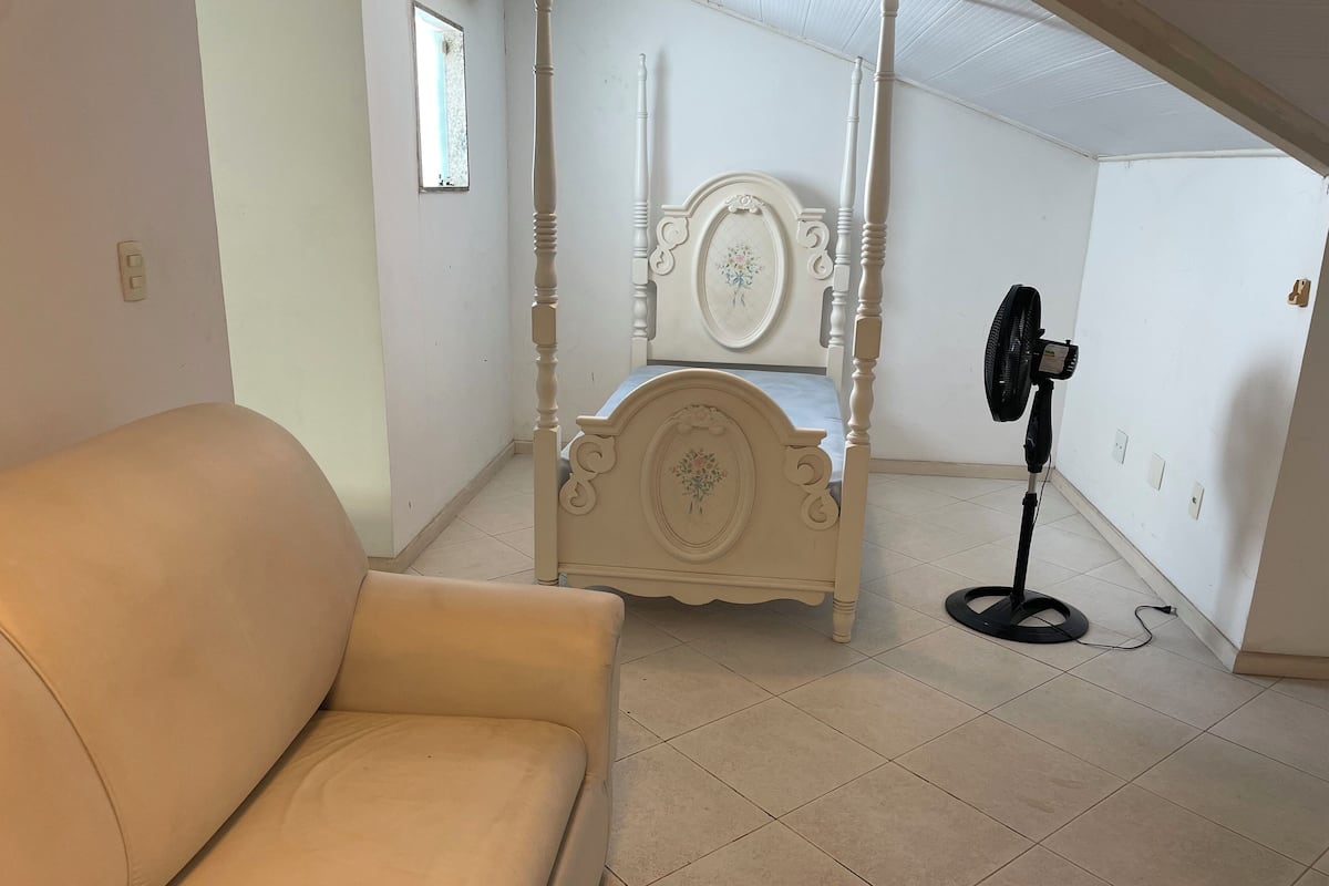 Una stanza con un divano beige, un letto a baldacchino bianco con ricami floreali, una ventola da terra e pavimento piastrellato.