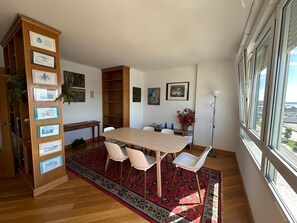 A dining area with a wooden table, white chairs, a red patterned rug, and a bookshelf with certificates.