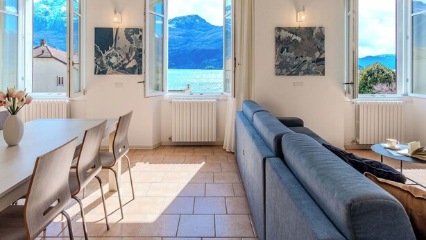 Una habitación con sofá, una mesa de comedor con sillas y vistas al mar y a las montañas a través de ventanas abiertas.