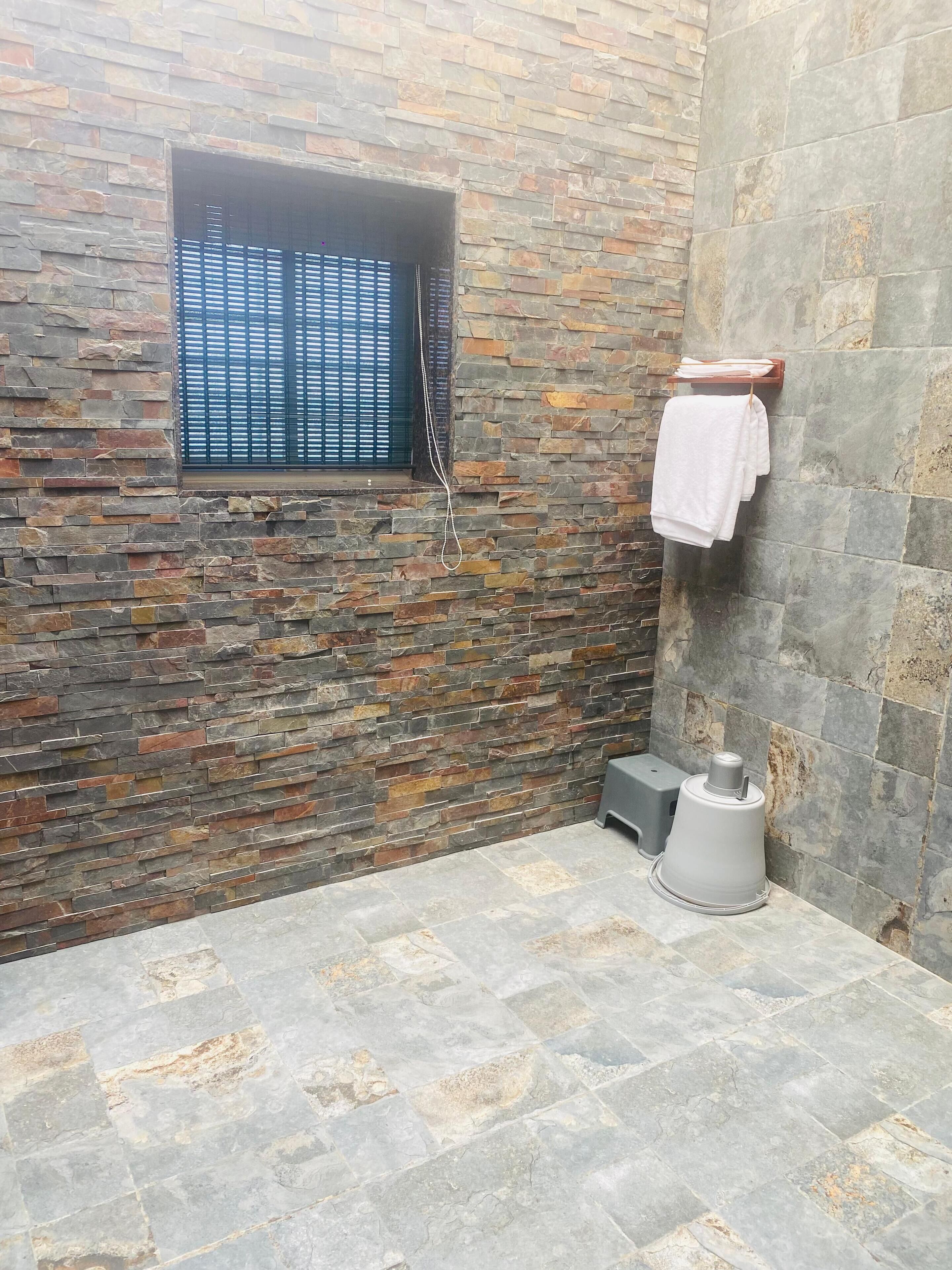 Un baño con pared de piedra, una ventana con persianas, un perchero con una toalla blanca y un dispensador de agua.