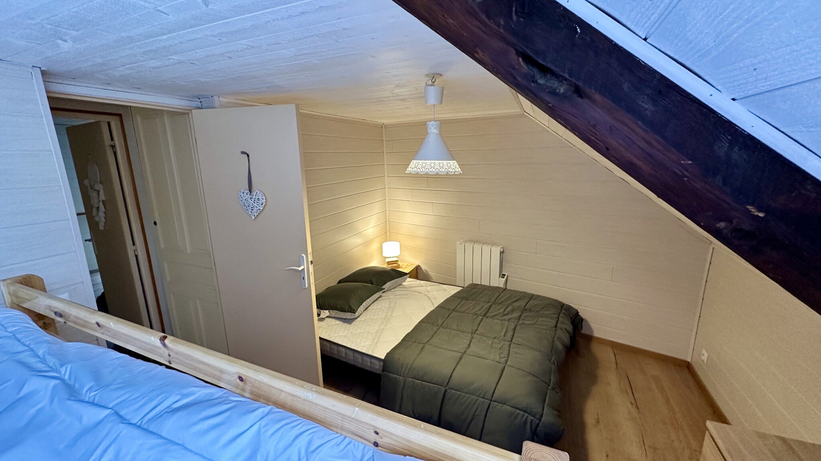 A small, cozy bedroom with a sloped ceiling, a single bed with green bedding, a bedside table with a lamp, and a decorative heart on the door.