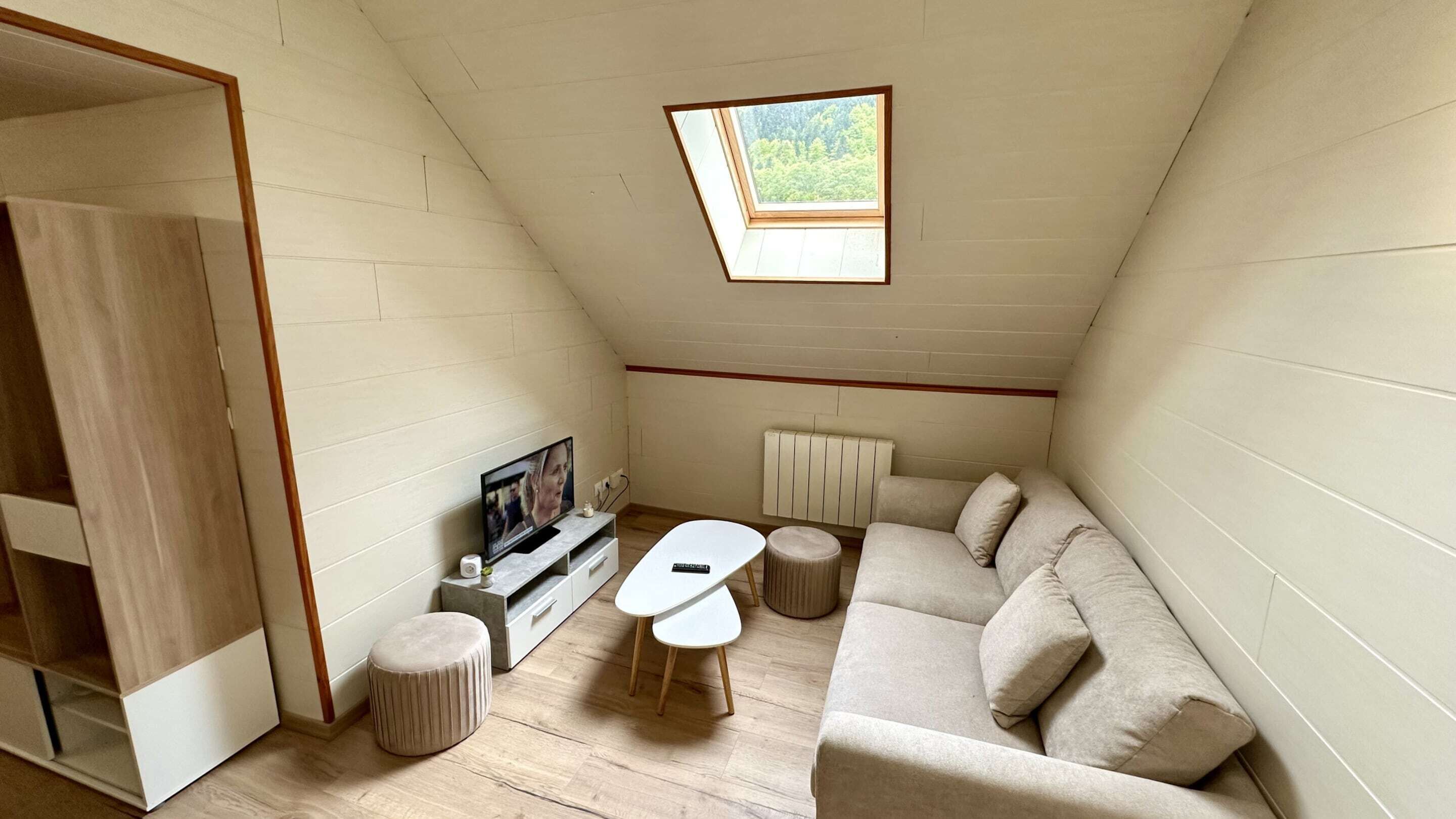 A small, triangular room with a sloped ceiling, a television, a sofa, two ottomans, and a radiator.