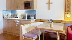 Eine kleine Küchenzeile mit Holzschränken, einer Mikrowelle und einem Spülbecken. An der Wand ist ein Kreuz angebracht, und es gibt eine Bank mit einer Blumenvase.