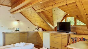 Une chambre confortable située dans les combles, avec un plafond en bois, un lit recouvert de draps blancs, une petite kitchenette et un téléviseur.