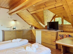 Ein gemütliches Dachzimmer mit Holzbalkendecke, einem Bett mit weißer Bettwäsche, einer kleinen Küchenzeile und einem Fernseher.