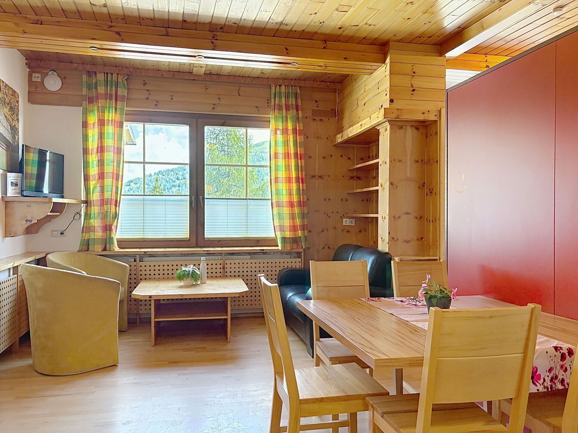 A cozy room with a wooden ceiling, a television on the wall, a sofa, a dining table with chairs, and a window with curtains.