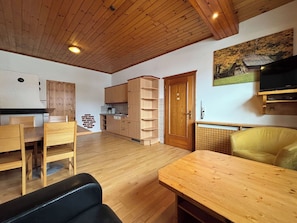 Ein kompakter Wohnbereich mit Holzboden, einer Küchenzeile mit Schränken und Spüle, einem Esstisch, einer Couch und einem an der Wand befestigten Fernseher.