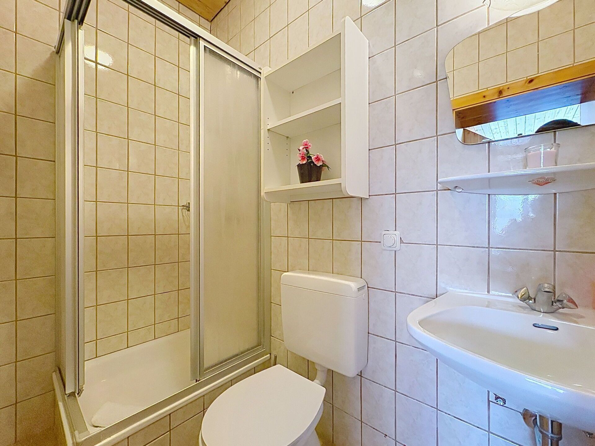 Et badeværelse med bruser, toilet, håndvask og hylder med toiletartikler.