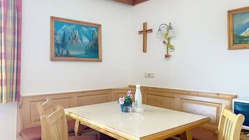 Een eetgedeelte met een houten tafel en stoelen, een kruis aan de muur en een schilderij van bergen.
