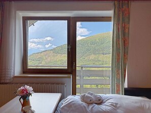 Ein Zimmer mit einem Bett, einem Fenster mit Blick auf einen bewaldeten Hügel, einem Heizkörper und einer Vase mit Blumen.