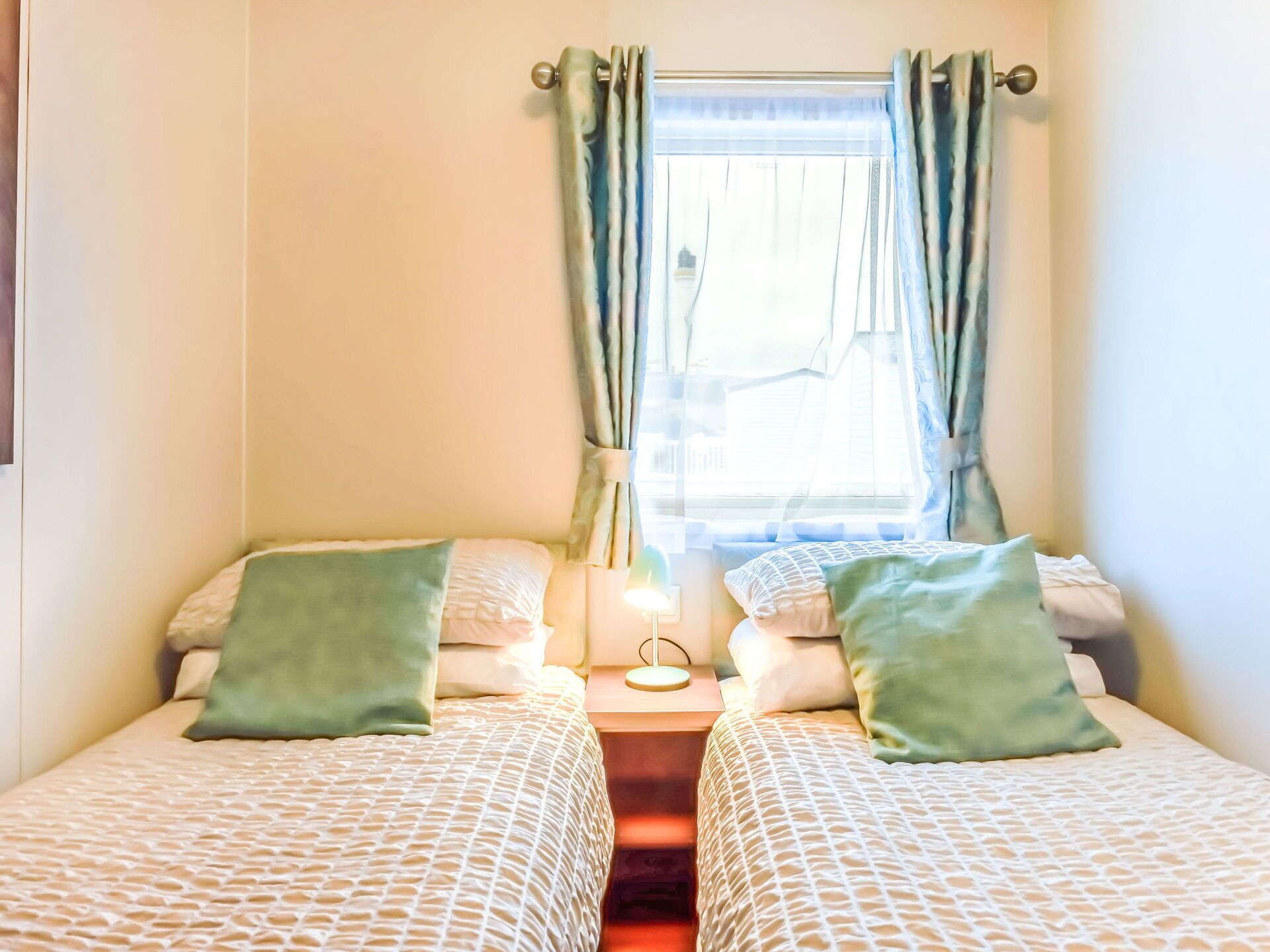 Une chambre avec deux lits, chacun agrémenté d’un oreiller vert et d’une couvre-lit à motifs. Une petite table de chevet avec une lampe se trouve entre les deux lits.