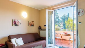 Ein gemütliches Wohnzimmer mit einem braunen Sofa, einem Glastisch gedeckt für das Essen und Blick auf einen Balkon mit Bäumen und blauem Himmel.