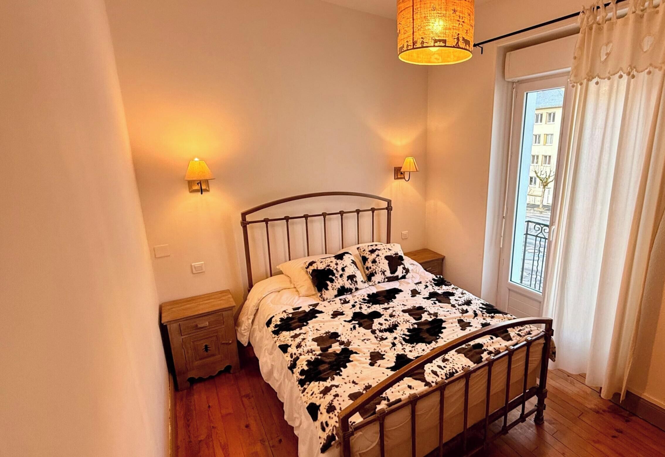 A bedroom with a metal bed frame, a cow print bedspread, two bedside lamps, a wooden nightstand, and a window with curtains.