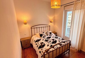 A bedroom with a metal bed frame, a cow print bedspread, two bedside lamps, a wooden nightstand, and a window with curtains. - L'abondance -Le Mont Dore 6pers (Mont-Dore)