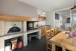 A modern kitchen with a built-in grill, wooden cabinets, and a dining area with chairs. - Stunning Villa With Pool in Sukosan (Sukosan)