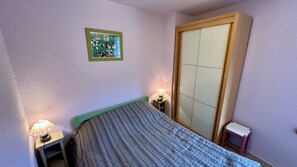 Une petite chambre à coucher comprenant un lit, deux lampes de chevet, une petite table, un mur orné d’un collage de photos encadrées et une armoire avec un miroir.