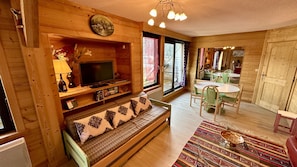 A cozy wooden cabin interior with a sofa, television, dining table, and chairs. - Sunny Sanctuary by the Town (Mont-Dore)